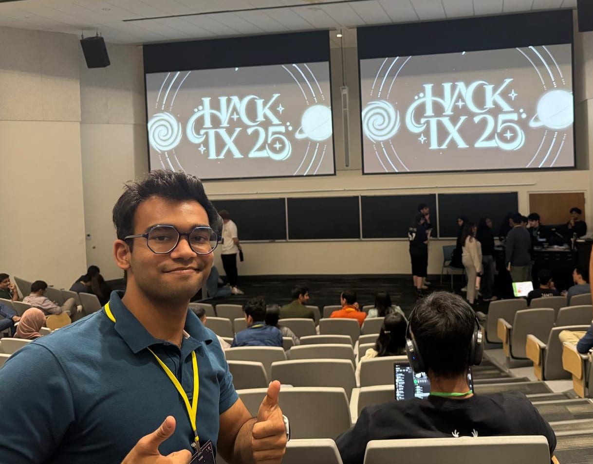 Manu in a lecture hall at HackTX 25, judge lanyard on, with the event title on both projection screens behind him.